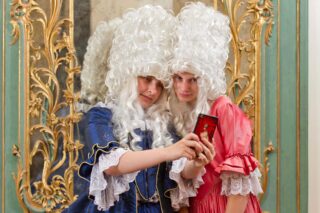 Zwei Personen in historischen Kostümen mit großen weißen Perücken machen ein Selfie vor einem dekorativen Spiegel. Die eine trägt ein blaues, die andere ein rotes Kleid.
