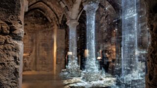 Ein altes historisches Gemäuer mit hohen >Säulen ist zu sehen. Darüber liegen helle Linien, die eine digitale Realität abbilden, die über den historischen Bauteilen liegt.