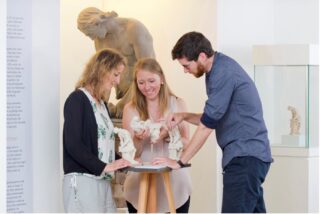 Drei Menschen stehen vor einer großen Skulptur und halten einen kleinen Entwurf in der Hand. Der Entwurf ist zum anfassen. er zeigt, wie man früher Skulpturen entworfen hat.