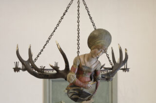 Ein Hängeleuchter mit einem Geweih und vorne einer Frauenfigur. Sie wird Lüsterweibchen genannt und stammt von dem meisterhaften Bildschnitzer Tilman Riemenschneider.