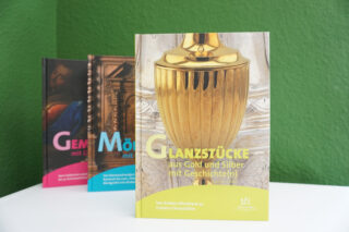 Ein aufgestelltes Buch mit dem Titel "Glanzstücke aus Gold und Silber mit Geschichten" auf einem weißen Tisch. Im Hintergrund sind weitere Bücher sichtbar, die in verschiedenen Farben gestaltet sind.