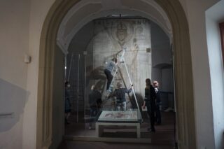 Ein Team von Menschen arbeitet an der Restaurierung des Kilianbanners. Eine Person steht auf einer Leiter, während andere die Arbeit beobachten. Im Vordergrund ist ein Glastisch zu sehen.
