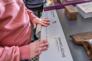 Ein Kind berührt eine braillebeschriftete Tafel mit dem Wort "Zahnfläche" in einer Ausstellung. Im Hintergrund sind verschiedene Materialien und Texturen sichtbar.