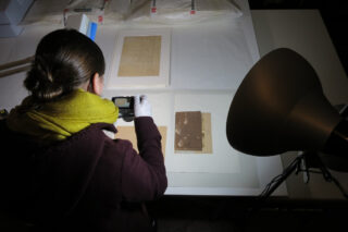 Eine Person mit einer Kamera, die vermutlich Dokumente oder Zeichnungen auf einem Tisch fotografiert. Im Hintergrund sind beleuchtete Unterlagen und eine Lampe sichtbar. Die Person trägt Handschuhe und einen bunten Schal.