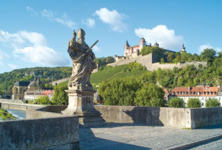 Eine Statue eines Mönchs steht im Vordergrund auf einer Brücke, mit Blick auf ein Schloss und Weinberg im Hintergrund. Der Himmel ist blau mit einigen Wolken und die Umgebung zeigt grüne Bäume und historische Gebäude.