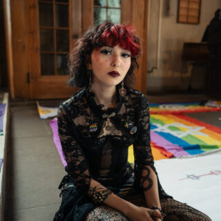 Eine dunkel gekleidete Frau mit rot-schwarzem Haar sitzt auf dem Boden vor einem bunten Regenbogenbild und Holzschiebetüren im Hintergrund.