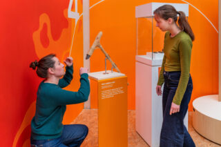 Zwei Frauen in einem Ausstellungsraum sehen sich ein Artefakt an, das auf einem orangefarbenen Sockel präsentiert wird. Eine Frau kniet und betrachtet das Artefakt, während die andere im Stehen interessiert zuschaut. Der Hintergrund ist mit kräftigen Farben gestaltet.