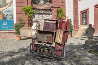 Ein Wagen mit kunstvoll gestapelten Holzkisten steht vor einem Museum. Der Wagen ist in einer schrägen Position und besteht aus verschiedenen Kisten in Rot- und Holztönen, während im Hintergrund eine Museumstür und Pflanzen sichtbar sind.