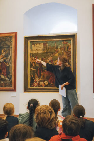 Eine Frau in einem schwarzen Oberteil steht vor einem großen Gemälde. Sie zeigt mit dem Finger auf das Bild und erklärt es einer Gruppe von Kindern, die aufmerksam sitzen. Im Hintergrund sind weitere Gemälde an der Wand sichtbar.