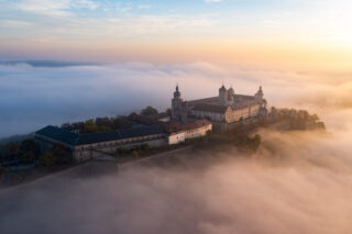 Eine Luftaufnahme einer beeindruckenden Burg, umgeben von Nebel, mit sanften Hügeln im Hintergrund. Der Himmel zeigt eine gemischte Farbpalette von sanften Orange- und Blautönen während des Sonnenaufgangs.